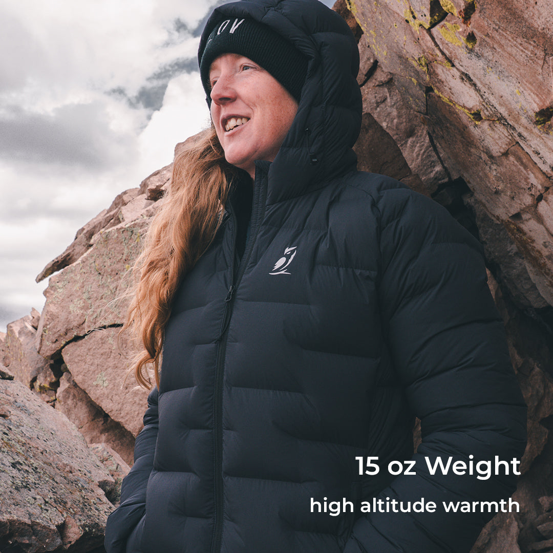 A woman wearing a black hooded puffer jacket and black knit beanie stands outdoors against large rock formations under a cloudy sky. White text in the lower right reads '15 oz Weight high altitude warmth.