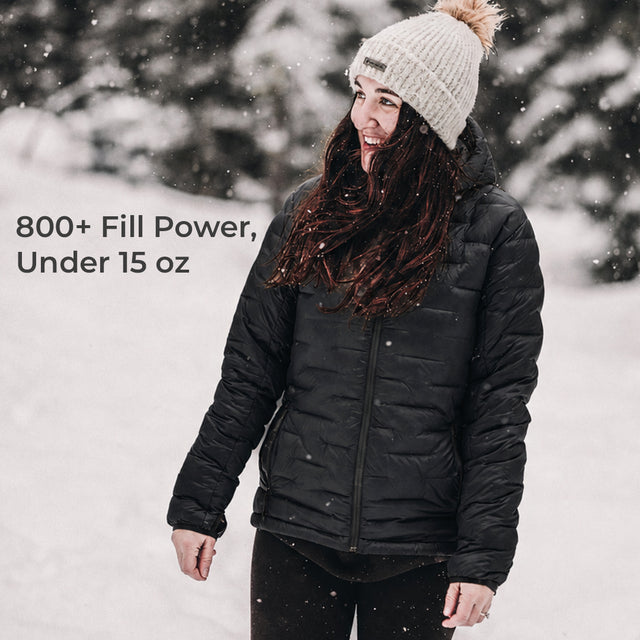 A woman wearing a black puffer jacket, black pants, and a cream knit beanie with a tan pom-pom stands in a snowy outdoor setting. Text on the left reads '800+ Fill Power, Under 15 oz'.