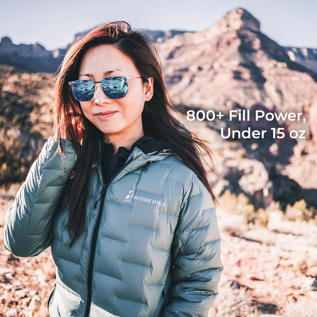 A woman wearing a light blue-gray puffer jacket and reflective sunglasses stands outdoors in a desert mountain landscape. White text on the right reads '800+ Fill Power, Under 15 oz.