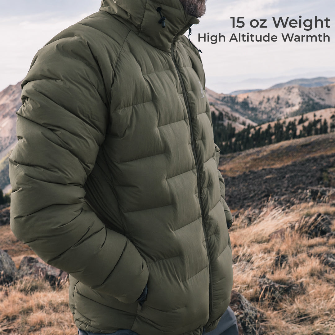 A man wearing a dark olive puffer jacket stands outdoors in a mountain landscape. Text in the upper right reads '15 oz Weight High Altitude Warmth'.