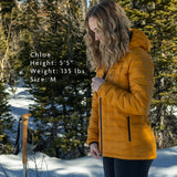 Woman wearing insulated hooded Zulu Down Jacket outdoors in snowy forest; model Chloe is 5'5", 135 lbs, wearing size medium for fit reference.