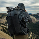 Hiker wearing a gray Outdoor Vitals Shadowlight backpack with gear strapped to the top and tripod stored in the side pocket, overlooking a rugged mountain ridgeline under cloudy skies.