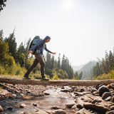 Backpacker balancing on a log over a mountain stream wearing the Altitude Sun Hoodie and backpack.
