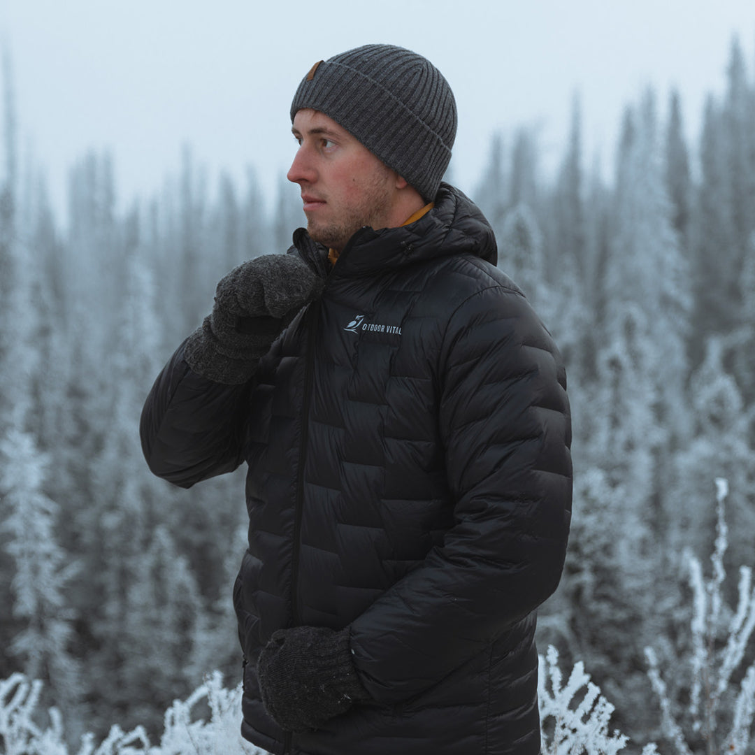 Person wearing a black NovaPro down insulated jacket and gray knit beanie, zipping the jacket up while standing in a snowy forest with frosted trees in the background.