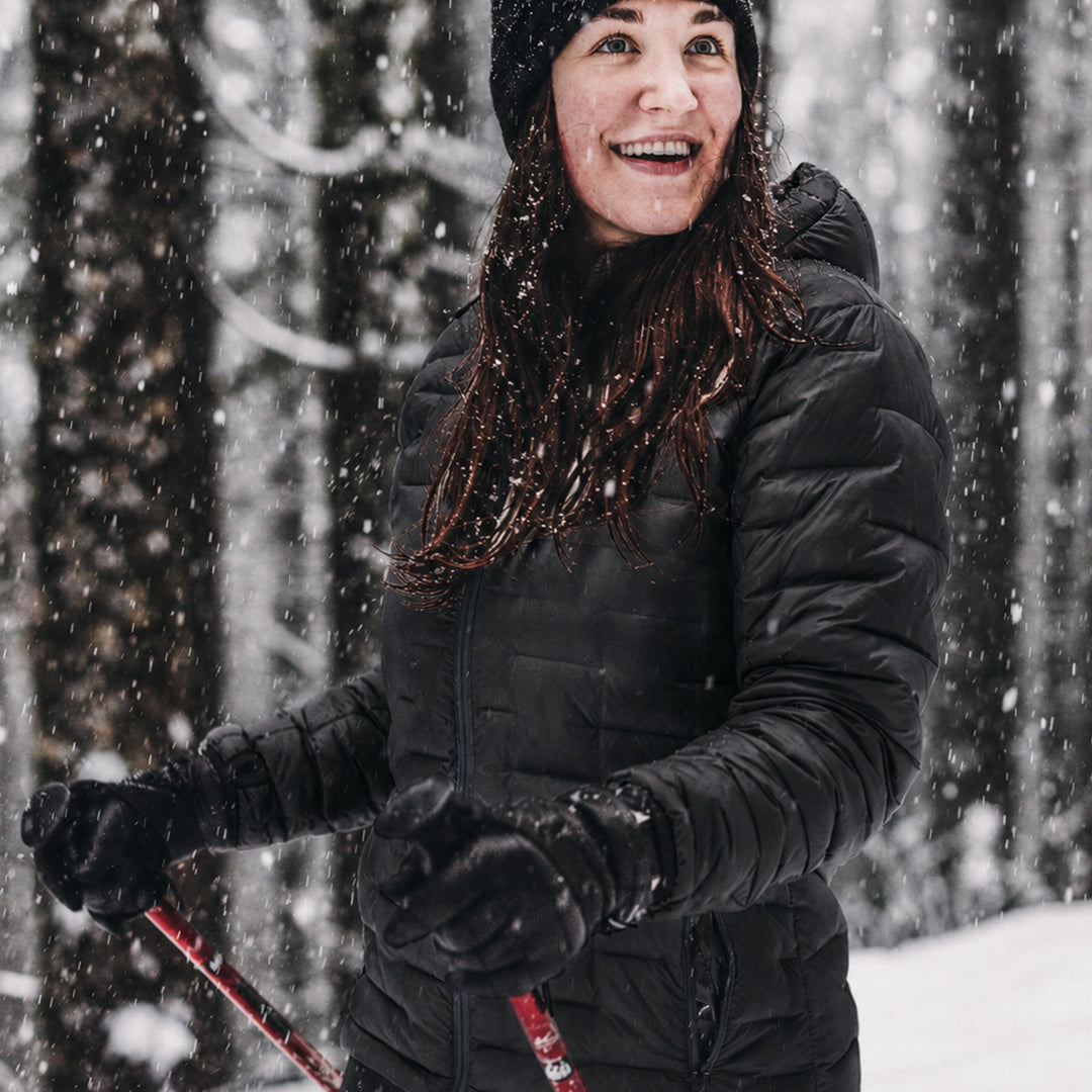 A woman smiling in a black puffer jacket, black beanie, and black gloves while skiing through falling snow in a wooded setting.