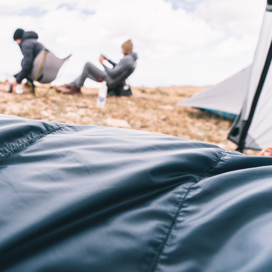 A close-up of a dark blue sleeping bag in the foreground, with a blurred outdoor camping scene in the background showing two people sitting near a tent on a rocky hillside.
