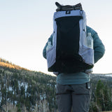 Hiker wearing Carbon Evo 50 backpack on mountain ridgeline with snow-covered forest in the background