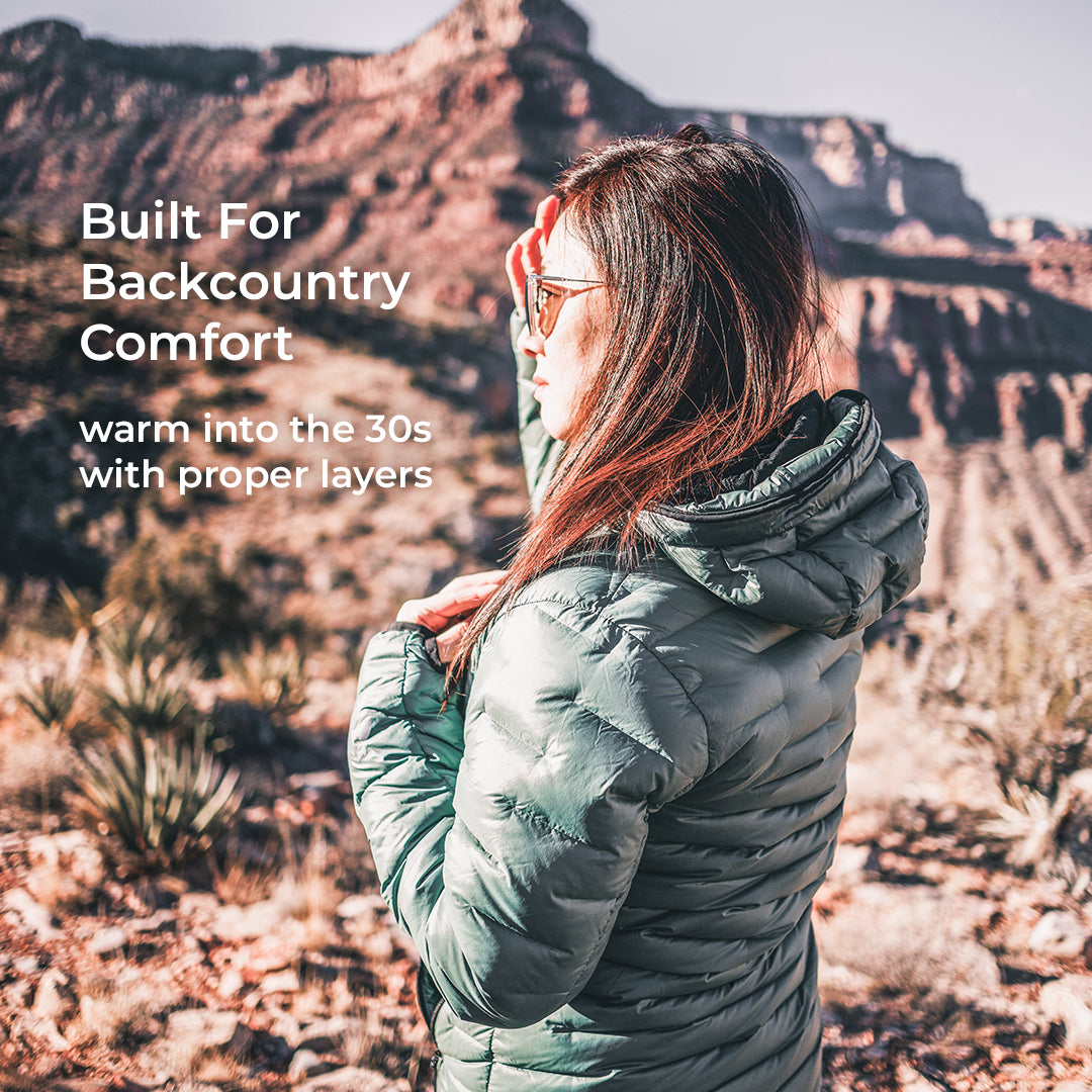 A woman wearing a sage green puffer jacket stands in a desert canyon landscape, looking out over the mountains. White text on the left reads 'Built For Backcountry Comfort' and 'warm into the 30s with proper layers'.