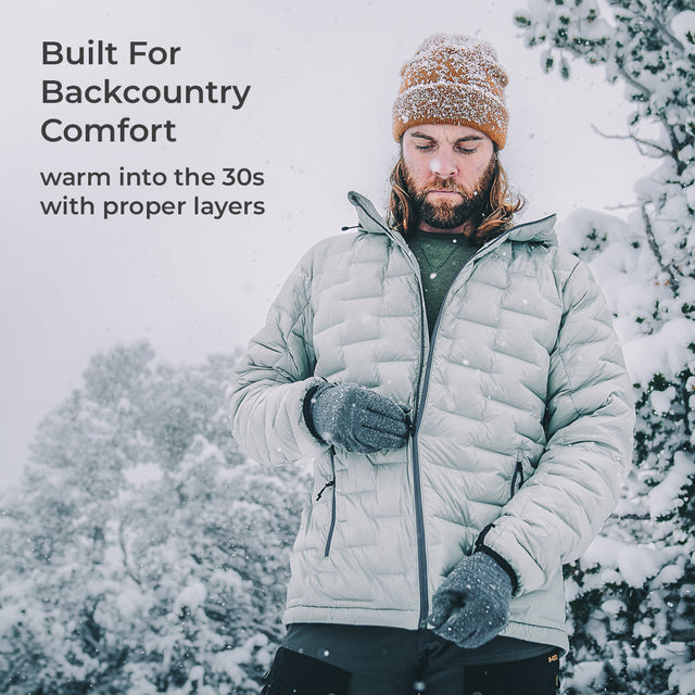 A man wearing a light gray quilted puffer jacket, brown knit beanie, and gray gloves stands in the snow with trees in the background. Text on the left reads: 'Built For Backcountry Comfort' and 'warm into the 30s with proper layers'.