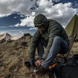 Backpacker wearing the Outdoor Vitals Vantage Alpine Down Jacket with hood up while cooking at a mountain campsite.