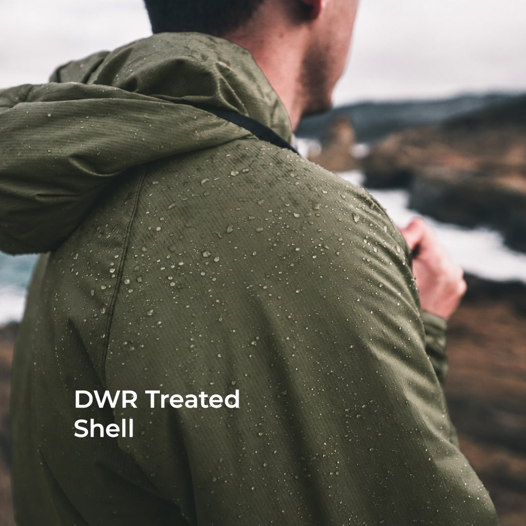A man viewed from behind wearing an olive green hooded jacket covered in water droplets, with a blurred outdoor coastal landscape in the background. White text on the lower left reads 'DWR Treated Shell'.