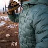 Close-up of a person wearing a green Outdoor Vitals insulated jacket and brown gloves, zipping the jacket up in a forested outdoor setting with patches of snow on the ground.