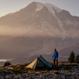 man stands by 2 person trekking pole tent pitched below rugged mountain