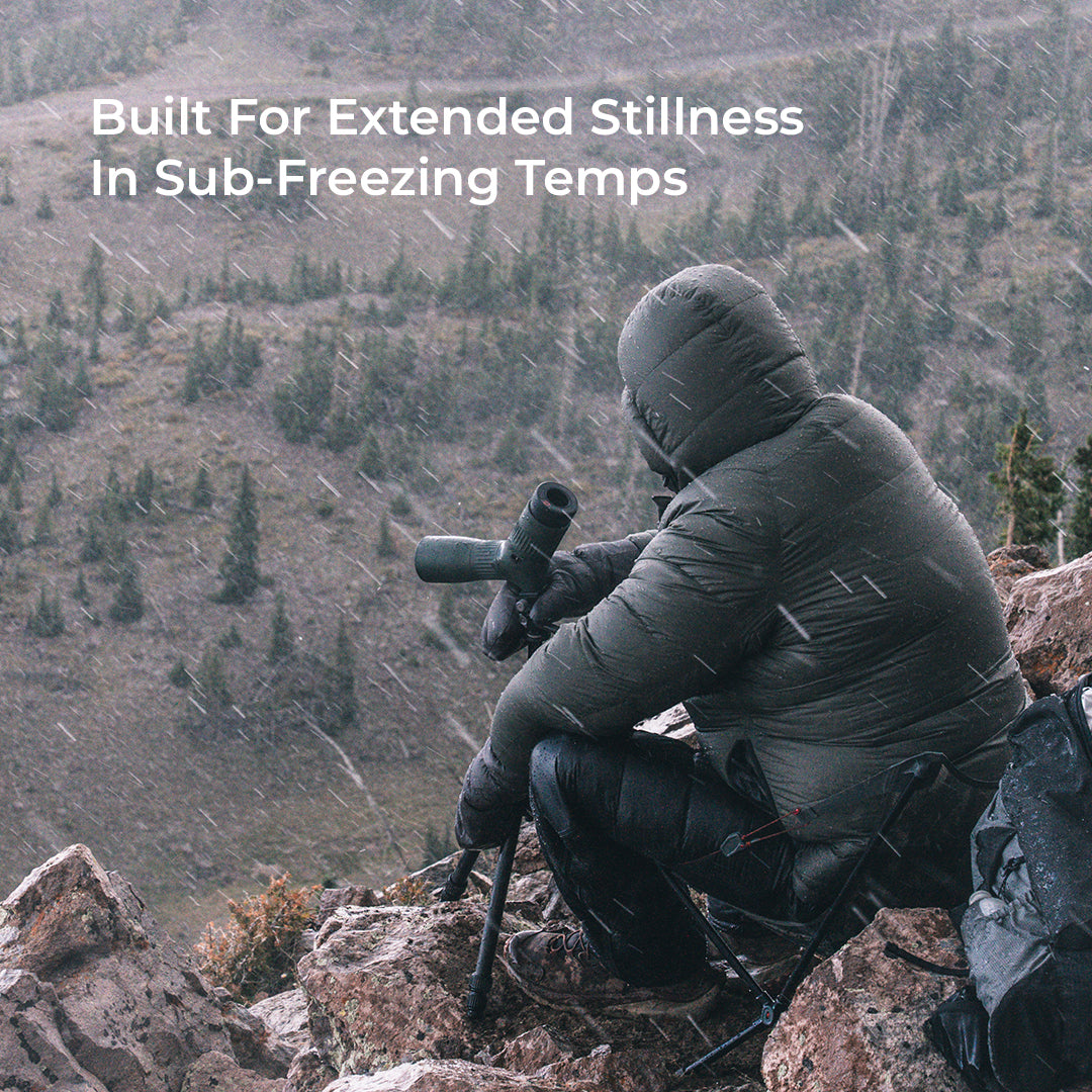 A person in a dark insulated hooded jacket crouches on a rocky mountainside in falling snow, using binoculars. Large white text at the top reads, 'Built For Extended Stillness In Sub-Freezing Temps.