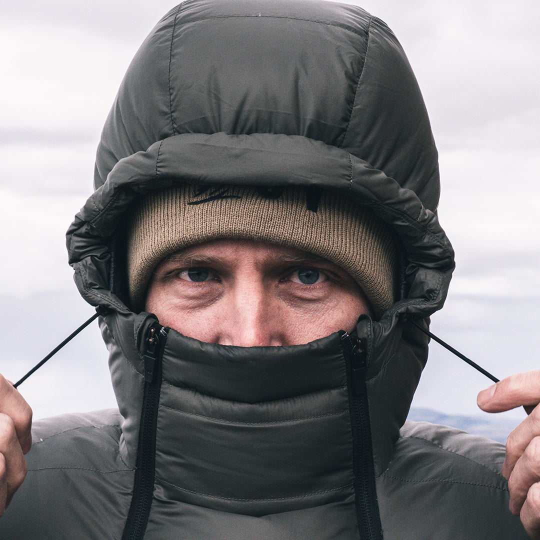 A close-up of a man wearing a dark puffer jacket with an integrated hood and face warmer, paired with a tan beanie, against an overcast sky.