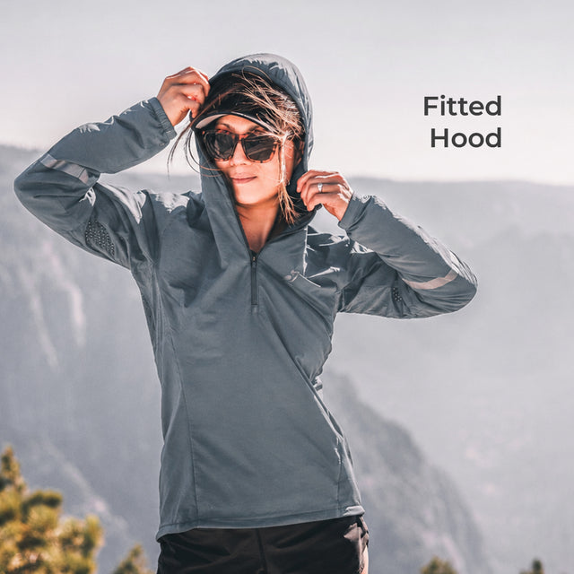 A woman wearing a fitted gray hooded quarter-zip athletic jacket stands outdoors with mountains in the background, adjusting the hood. The text 'Fitted Hood' appears in the upper right.