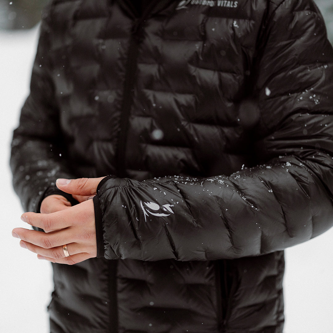 Close-up of a person wearing a black Outdoor Vitals insulated jacket in the snow, showing thumb loop cuffs with the brand’s logo on the sleeve.