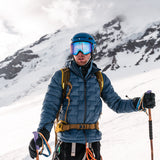 Mountaineer wearing a blue Outdoor Vitals insulated jacket, helmet, and ski goggles, holding trekking poles while standing on a snowy mountain slope with rugged peaks in the background.