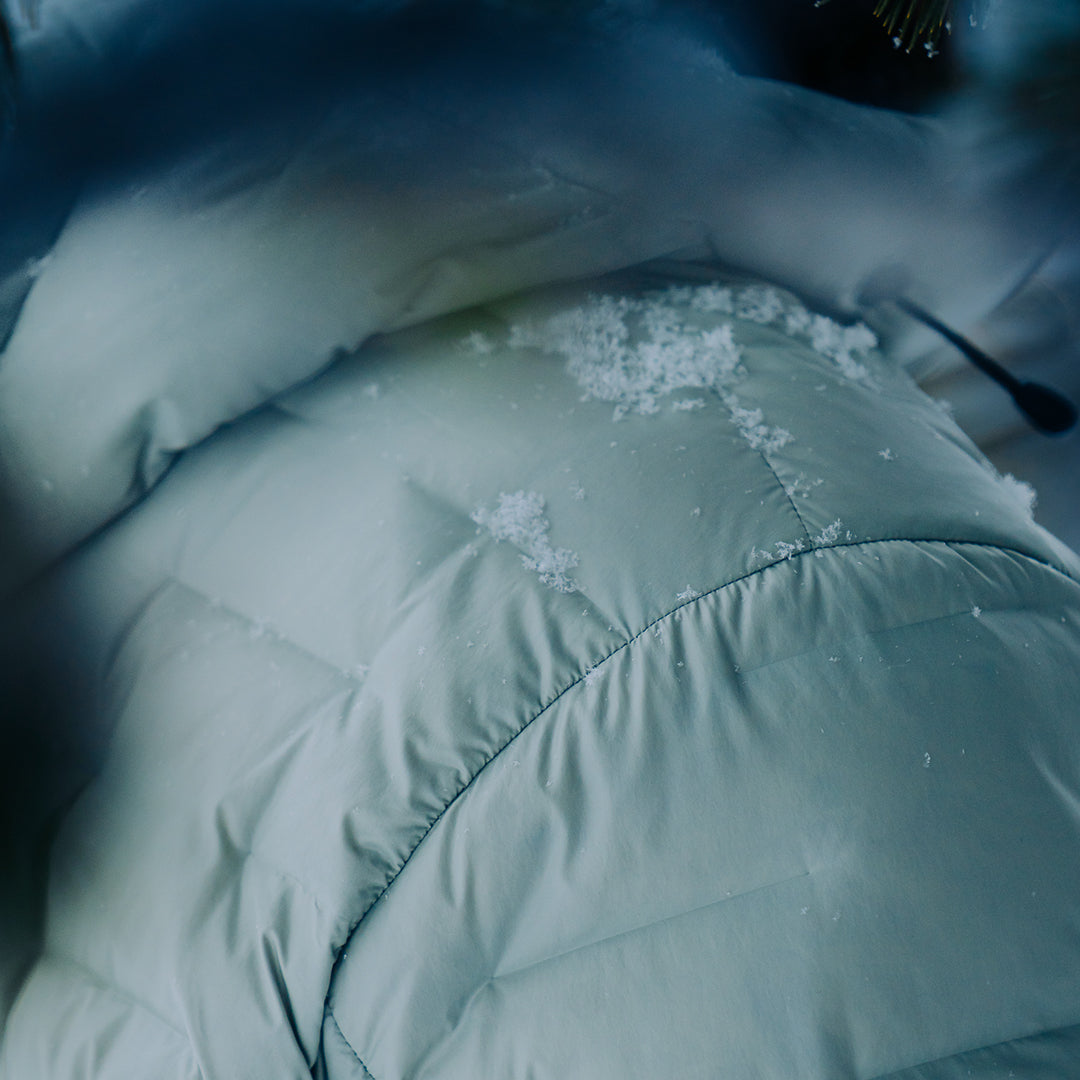 Close-up of a light gray Outdoor Vitals insulated jacket with snow resting on the shoulder and hood, showing the fabric’s weather resistance.
