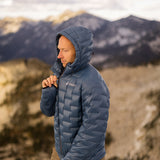 Person wearing a blue Outdoor Vitals NovaPro insulated jacket with the hood up, zipping it closed while standing in a mountain landscape.