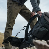 Close side view of man wearing green/brown hiking pants while picking up a backpack