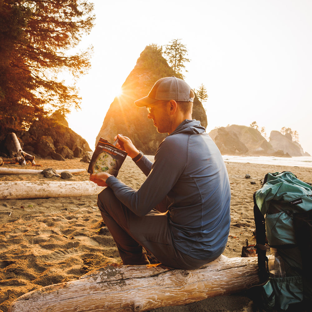 side view of hiker with blue sun hoodie eating dinner on log
