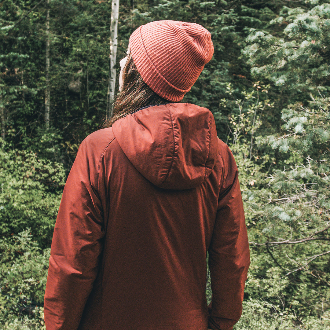 A person viewed from behind wearing a rust-colored hooded jacket and a matching ribbed knit beanie, standing in a dense green forest.