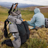 Rear view of Outdoor Vitals Shadowlight backpack on the ground while hiker wearing a blue jacket sits down on an open alpine landscape.