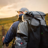 Back view of a hiker wearing the Outdoor Vitals Shadowlight backpack, carrying a rolled sleeping pad secured on top, hiking at golden hour with water bottle visible in side pocket.