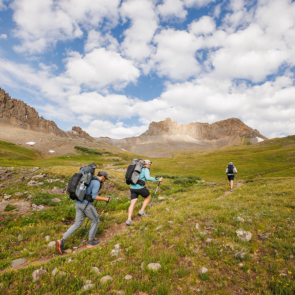 Three hikers carrying Outdoor Vitals Shadowlight backpacks trekking uphill on a trail through a green alpine meadow with rocky peaks in the background.