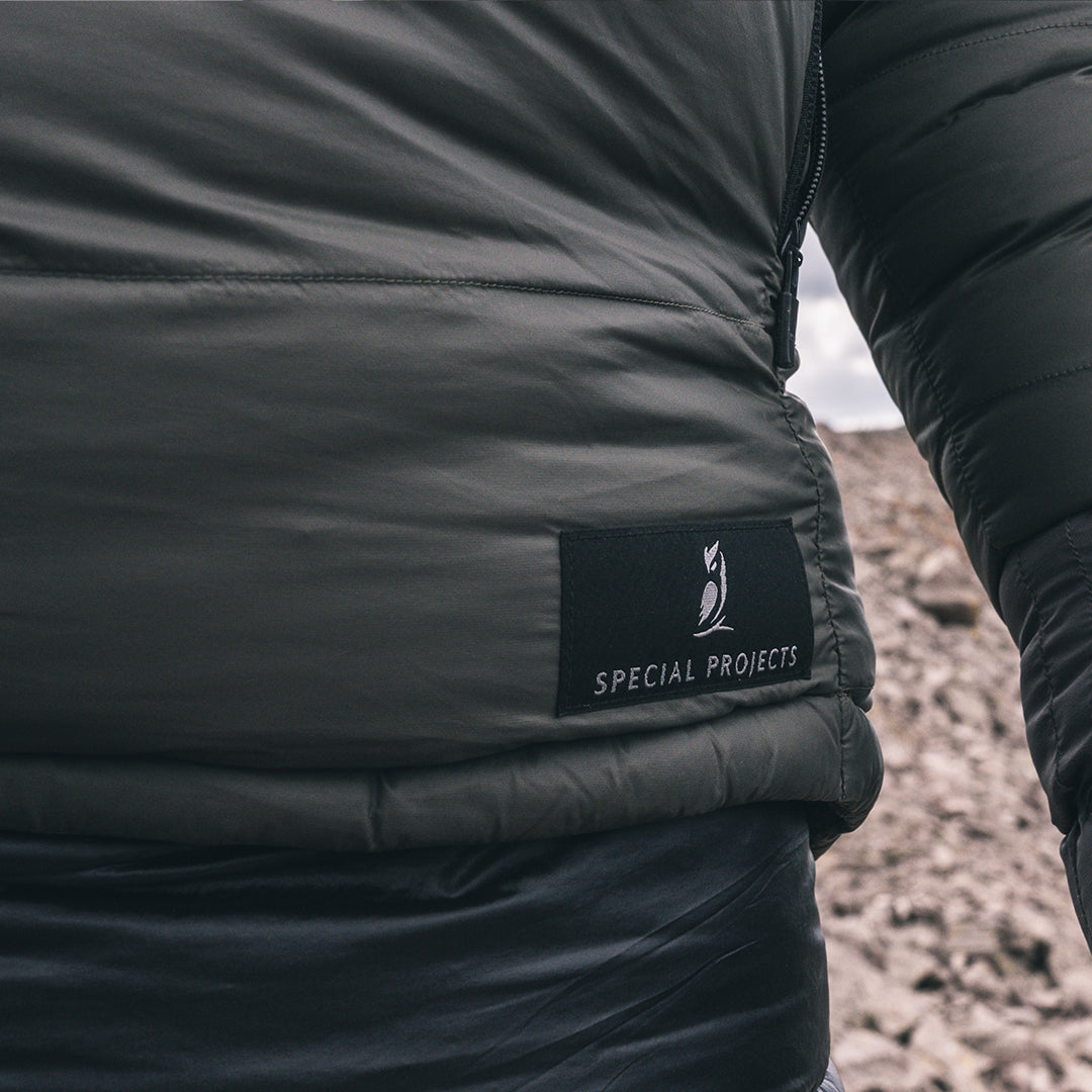 Close-up of a person wearing a dark gray puffer jacket, showing a black brand patch that reads 'SPECIAL PROJECTS' on the side, with a rocky outdoor background.