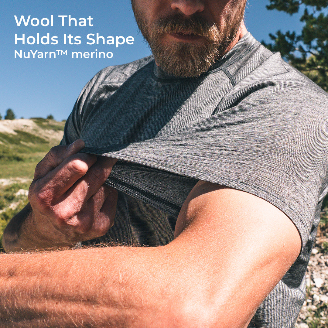 A close-up outdoor shot of a bearded man lifting his short-sleeve shirt to show the fabric shape retention and stretch. Text in the top left reads 'Wool That Holds Its Shape NuYarn™ merino'.