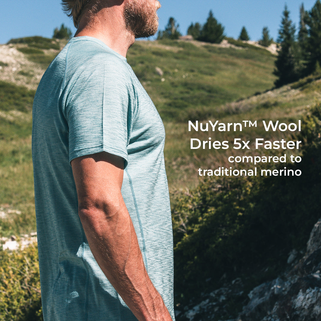 A man wearing a light heathered short-sleeve t-shirt stands outdoors on a grassy mountain slope, with white text on the right reading 'NuYarn™ Wool Dries 5x Faster compared to traditional merino'.