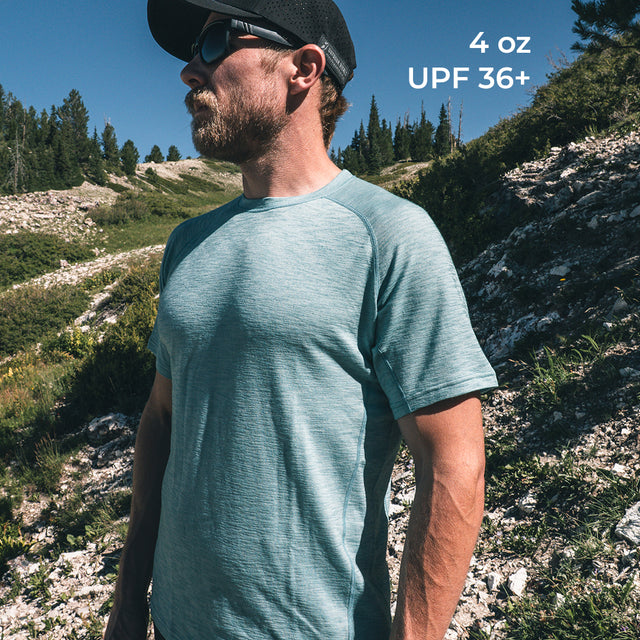 A man wearing a short-sleeve athletic t-shirt with a black cap and sunglasses, standing outdoors in a rocky, grassy mountain environment under a clear blue sky. White text in the top right corner reads '4 oz UPF 36+'.