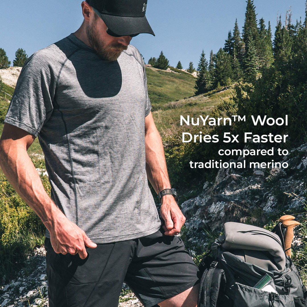 A man wearing a wool short-sleeve athletic t-shirt and black shorts stands outdoors on a rocky mountain path next to a hiking backpack. White text on the right reads 'NuYarn Wool Dries 5x Faster compared to traditional merino'.
