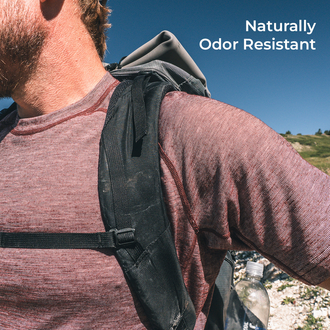 A close-up outdoor shot of a person wearing a backpack over a long-sleeve shirt, with blue sky and a rocky hillside in the background. White text in the top right reads 'Naturally Odor Resistant'.