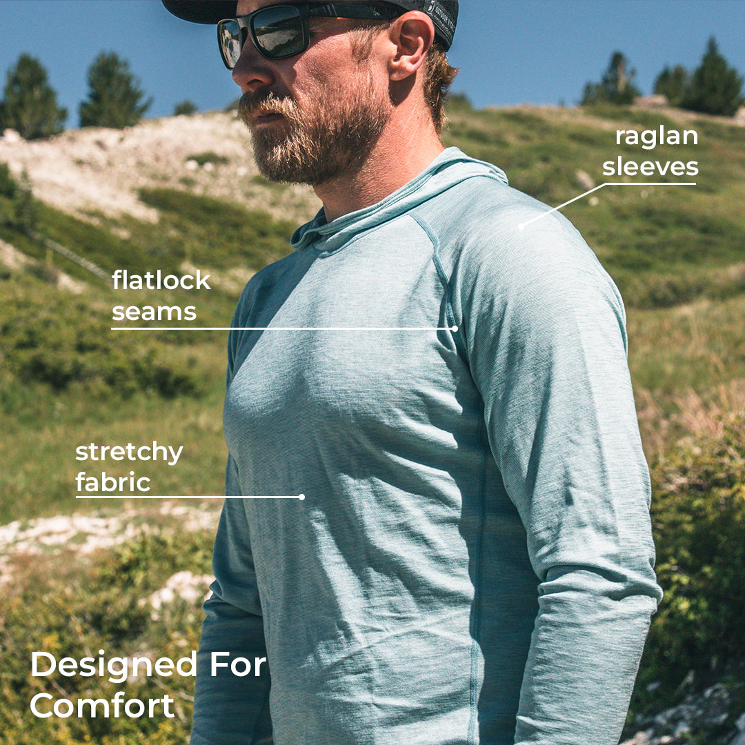A man wearing a long-sleeve performance hoodie stands outdoors on a grassy hillside. White text labels point to features of the garment: 'flatlock seams,' 'stretchy fabric,' 'raglan sleeves,' and 'Designed For Comfort.