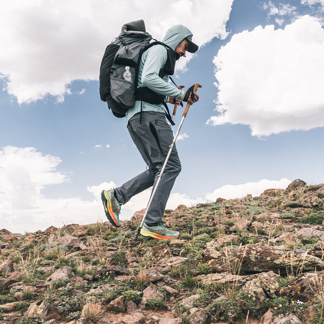 A hiker wearing a light merino hoodie, hiking pants, and a backpack with a water bottle, walking up a rocky mountain slope with a trekking pole under a blue sky with white clouds.