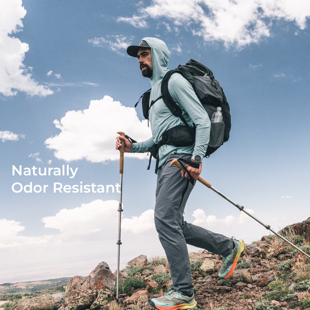 A man hiking on a rocky mountain under a blue sky with clouds, wearing a hooded long-sleeve shirt, hiking pants, and carrying a black backpack with trekking poles. White text on the left reads 'Naturally Odor Resistant'.