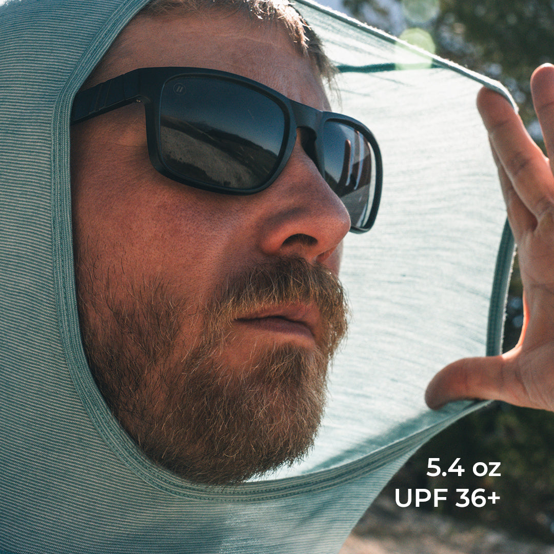 Close-up of a man wearing a light wool hooded sun shield and black sunglasses outdoors, with text in the lower right reading '5.4 oz UPF 36+'.