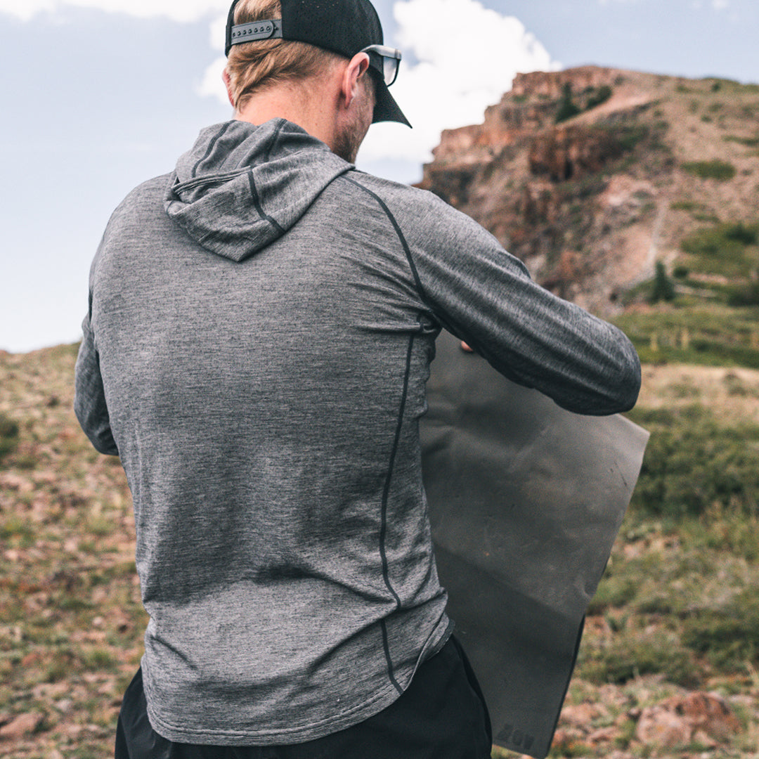 A man wearing a dark gray heathered long-sleeve merino wool hoodie, viewed from behind as he holds a gray foam pad in an outdoor mountain setting.