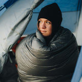 close view of a female hiker wrapped in quilt looking out her tent door