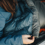 Close-up of a person in a blue jacket holding the edge of a puffy down top quilt, showing the insulated neck baffle and stitching details.