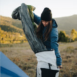 Camper wearing a blue jacket and black beanie pulling a gray top quilt out of a white backpack in a grassy mountain setting during sunrise.