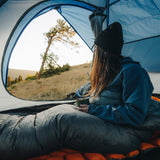 Camper sitting inside a tent in a down top quilt and blue jacket, holding a cup and looking out at a grassy hillside with trees during sunrise or sunset.