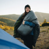 Smiling backpacker in a blue jacket and black beanie draping a gray top quilt or sleeping bag over their shoulders while standing outdoors in a mountain meadow at sunrise.