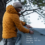 Man wearing insulated hooded down jacket while setting up a tent outdoors; model Tyler is 5'11", 165 lbs, wearing size medium for fit reference.