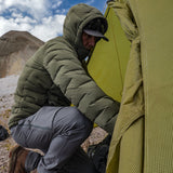 Man wearing Vantage Alpine Down Jacket setting up tent in the mountains – Outdoor Vitals Vantage down jacket in olive green, providing ultralight warmth and protection while pitching a tent in alpine conditions.