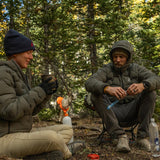 Campers staying warm in Outdoor Vitals Vantage Alpine Down Jackets, enjoying hot drinks at a mountain campsite with pack goat in the background.