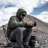 Hiker sitting on a camp chair wearing the Outdoor Vitals Vantage Alpine Down Jacket with hood up in alpine terrain.